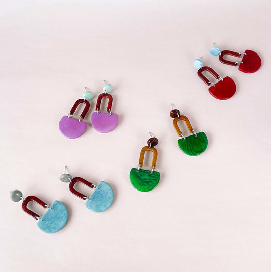 Six pairs of colorful stud earrings arranged on a pale pink background.