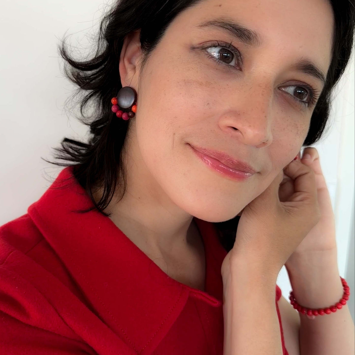 A close-up portrait of a woman with dark hair wearing a bright red top and matching red beaded jewelry, including a red bracelet and a red and black beaded earring, against a plain light background.