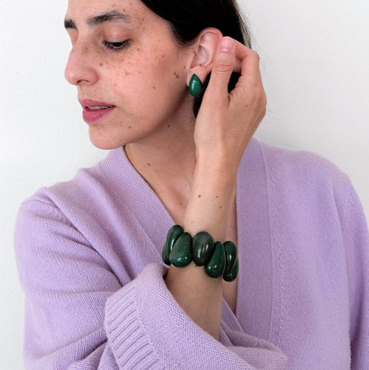 A woman wearing a lavender knit sweater and a dark green beaded bracelet, adjusting a matching dark green beaded earring on her ear against a plain white background.