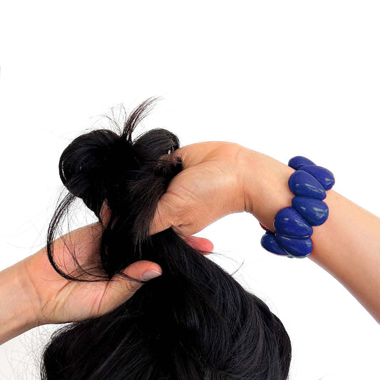 A close-up of a person's hands gathering and securing a section of long black hair with a small black hair tie, against a plain white background. The person is also wearing a blue beaded bracelet.