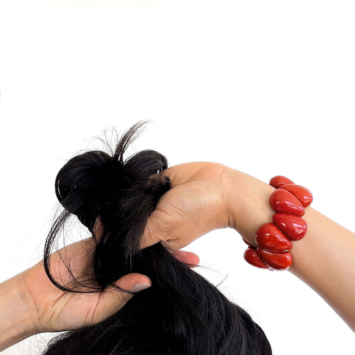 A close-up of a person's hands gathering and tying a high ponytail with long, straight black hair. The person is wearing a bright rust beaded Tagua Nut bracelet on one wrist, set against a plain white background.