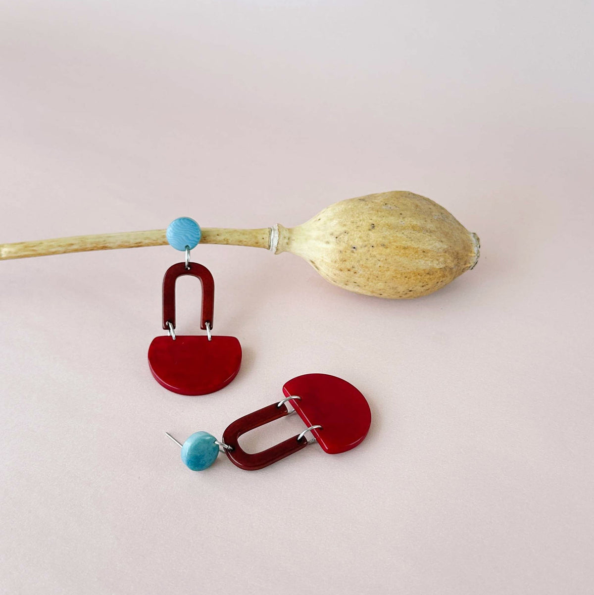 A pair of red geometric statement earrings with small turquoise blue bead accents, displayed on a light pink background with a dried seed pod prop.