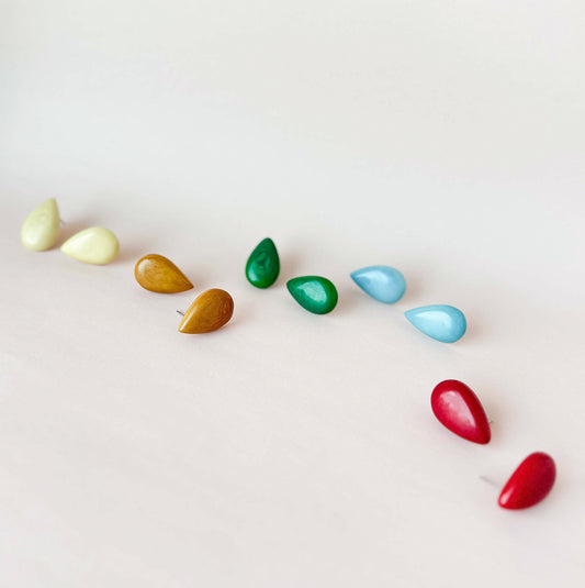 A set of small teardrop-shaped tagua nut stud earrings in multiple colors, including sand, mustard, emerald green, sky and red, arranged in a diagonal line on a plain off-white background.