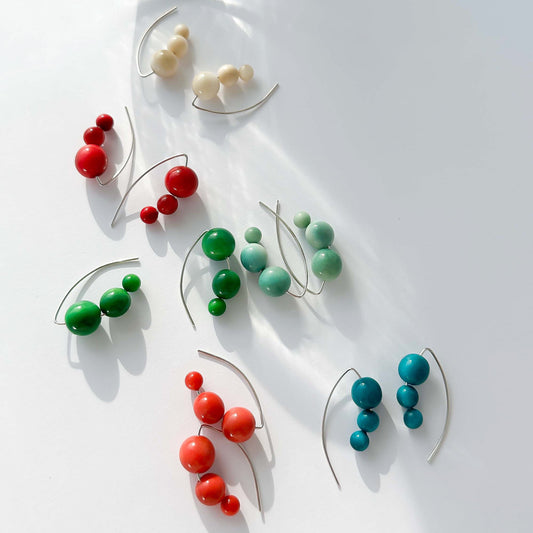 A set of colorful beaded wire earrings arranged on a white surface with a clear ribbed glass tumbler at the top. The earrings feature clusters of red, green, mint green, and teal beads on thin silver-tone wires.