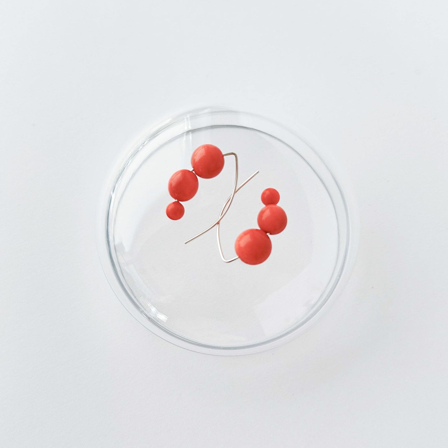 A minimalist wire-frame jewelry piece featuring five glossy red spherical beads on a thin gold-toned branch, displayed inside a clear glass dome against a clean white background.