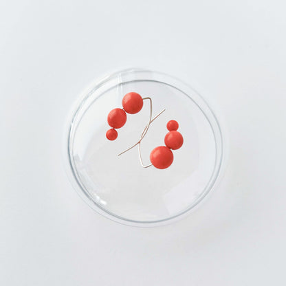 A minimalist wire-frame jewelry piece featuring five glossy red spherical beads on a thin gold-toned branch, displayed inside a clear glass dome against a clean white background.