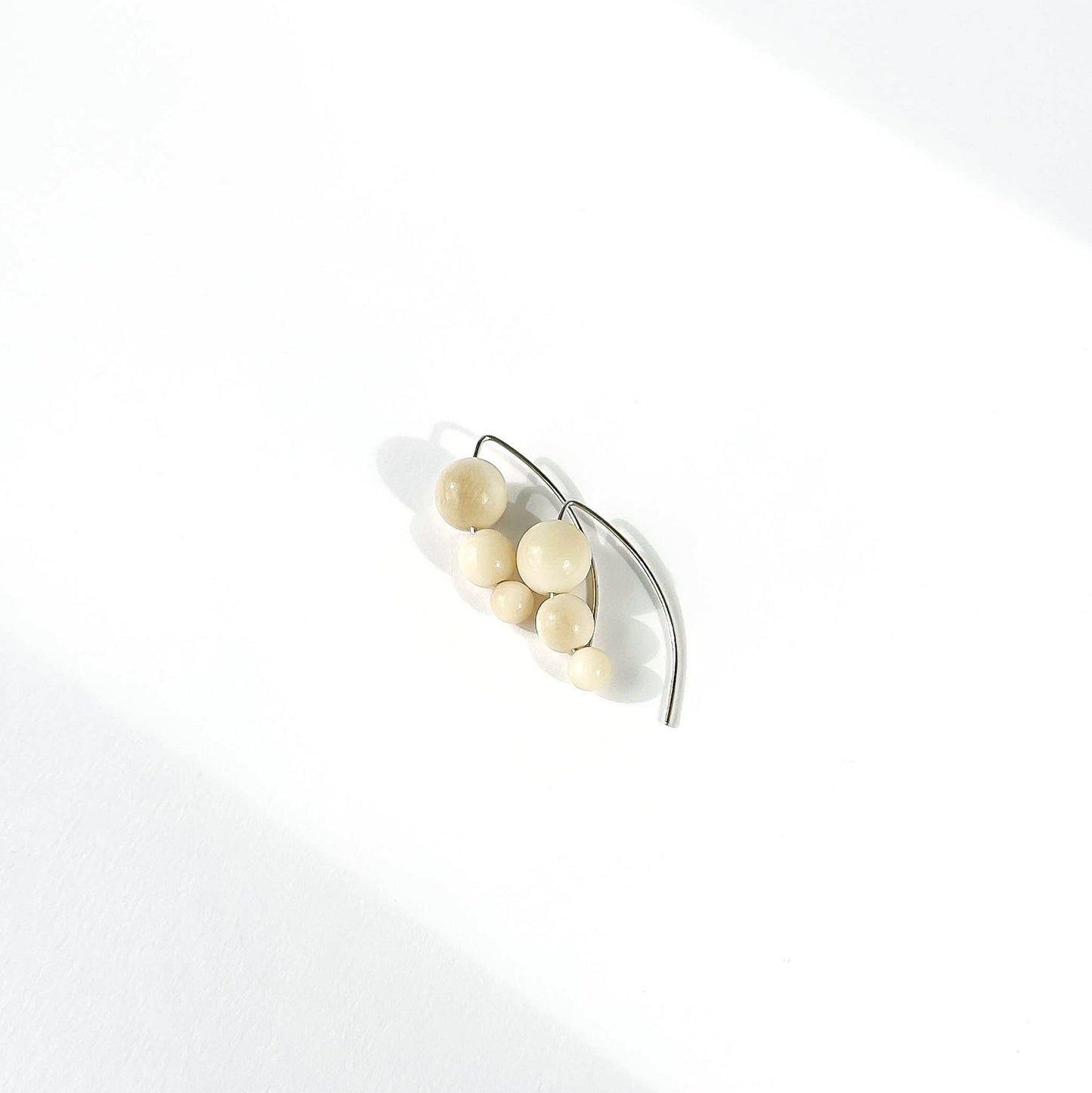 A pair of minimalist silver earrings featuring a cluster of small, matte cream-colored beads on a curved silver hook, photographed on a clean white surface with soft diagonal shadows.