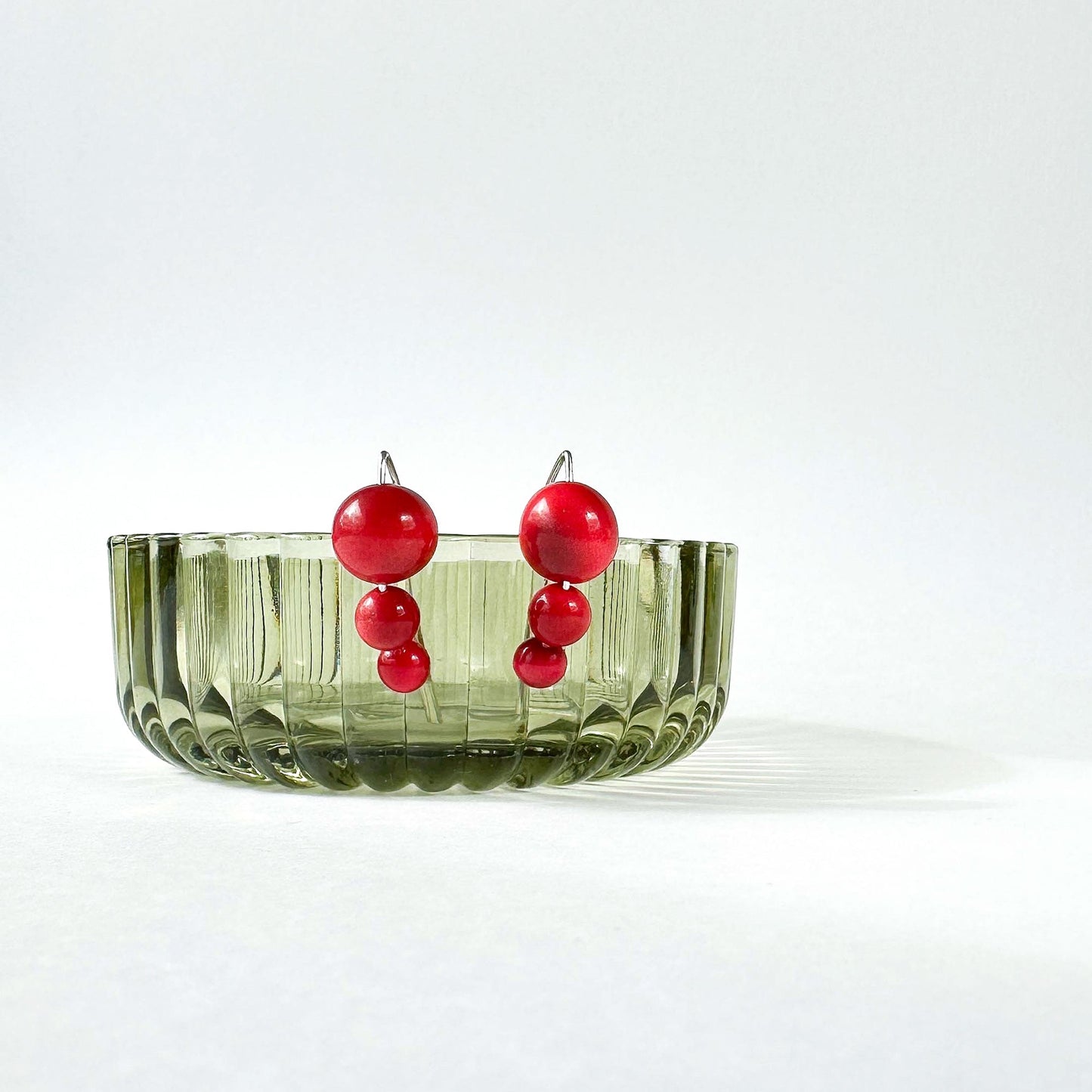 A pair of red beaded drop earrings with silver-toned posts, displayed in a ribbed translucent olive green glass bowl against a clean white background.