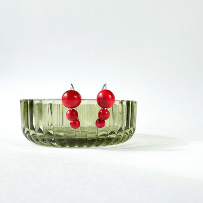 A pair of red beaded drop earrings with silver-toned posts, displayed in a ribbed translucent olive green glass bowl against a clean white background.